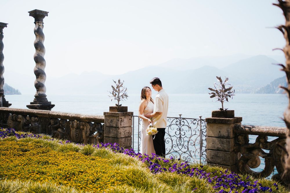 A romantic couple embraces on a hilltop terrace with ornate stone pillars and colorful flowers overlooking misty mountains.