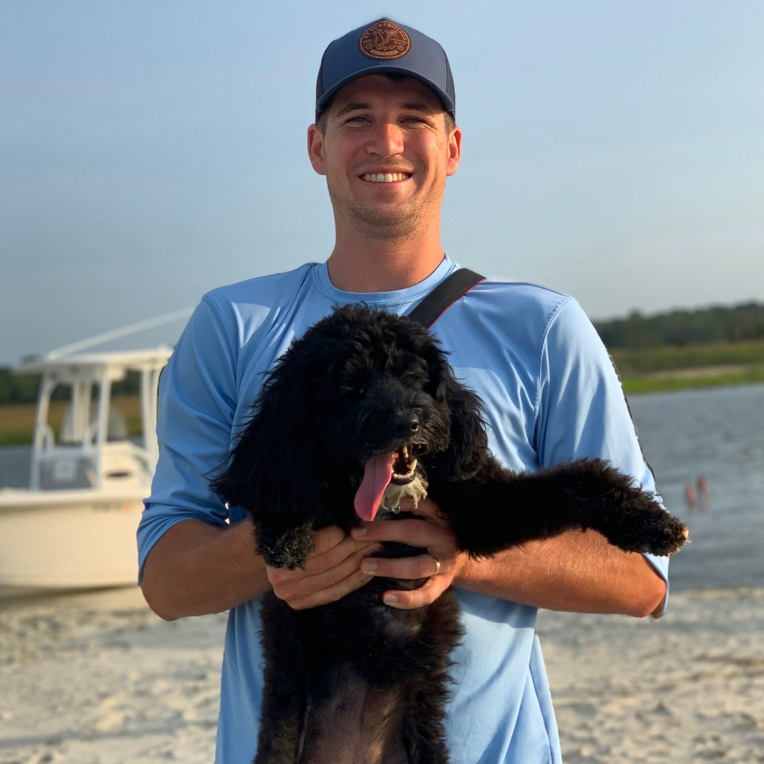 Person in blue shirt holding a happy black dog on a beach with boat in background.