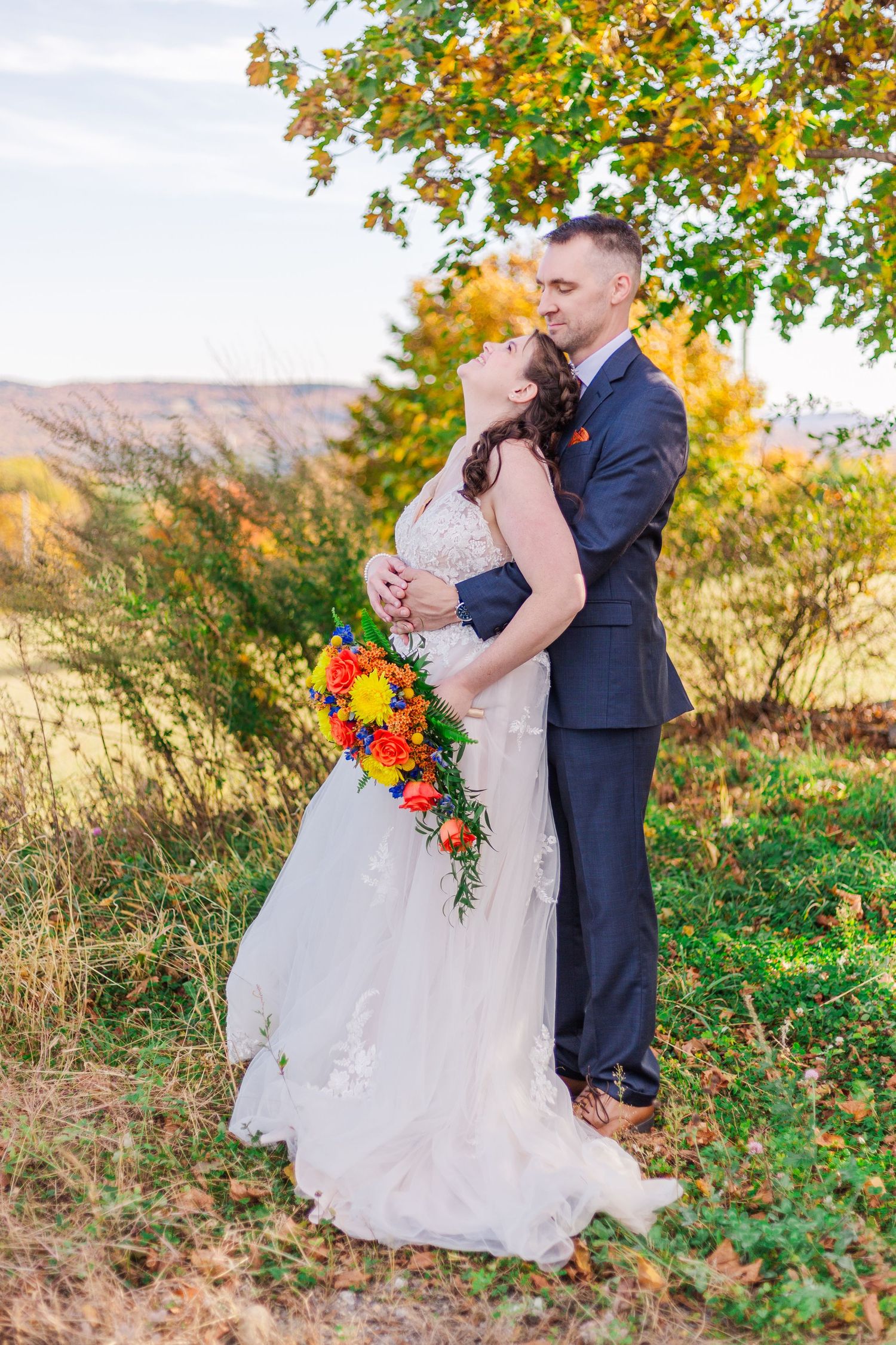 Bride and groom embrace in romantic fall setting with colorful bouquet and autumn trees.