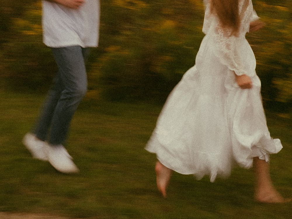 Blurred motion shot of a couple running on grass, woman in white dress, man in jeans and white shoes.
