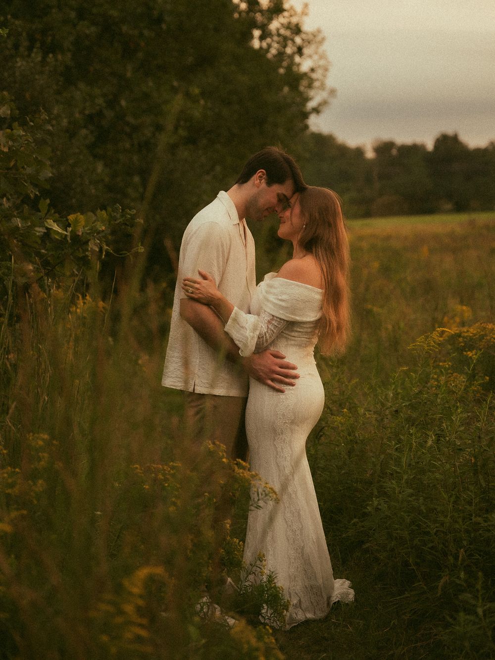 Couple embracing in a lush green field at golden hour, woman in white lace off-shoulder gown, romantic outdoor portrait.
