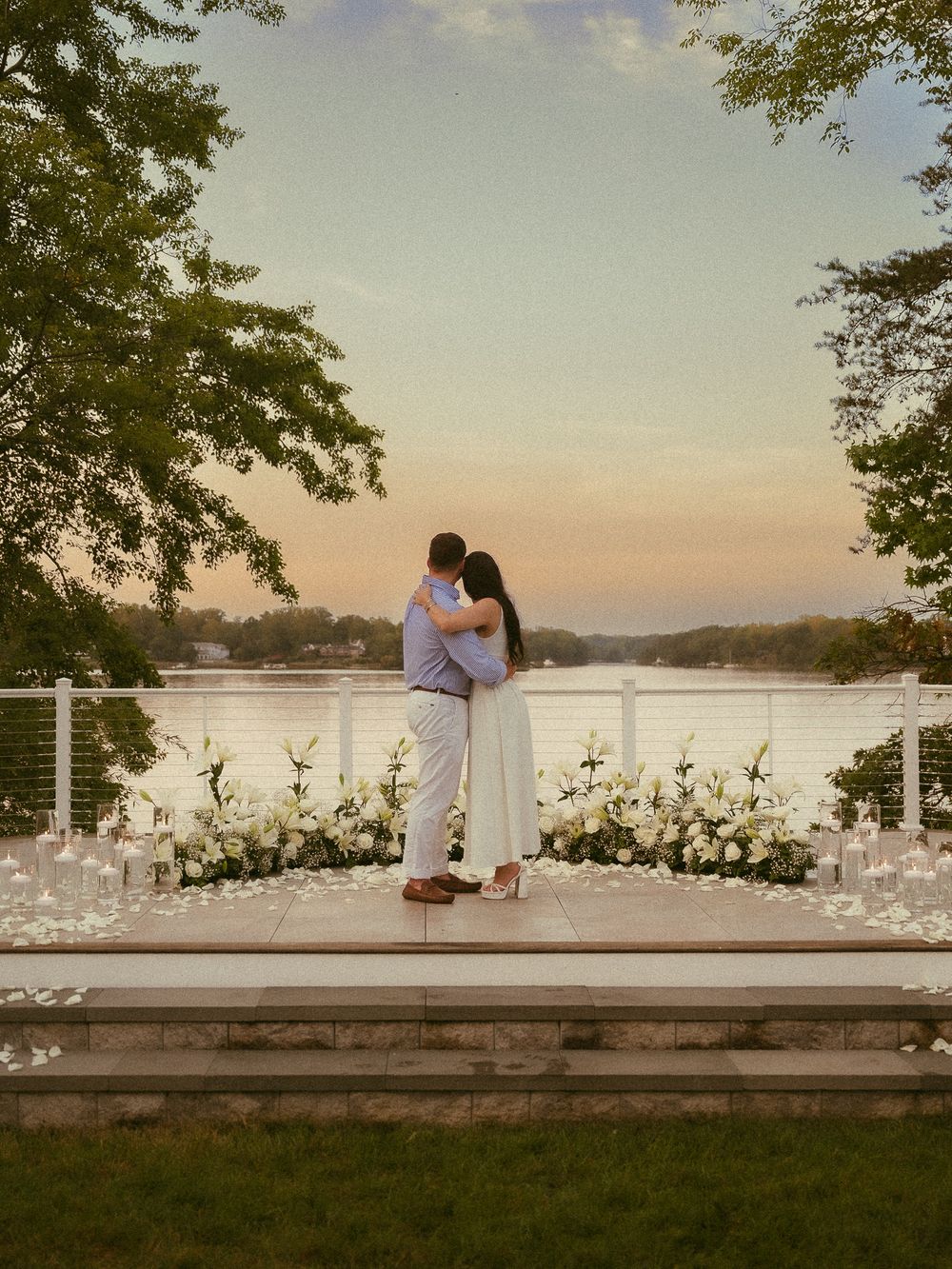 Couple embracing on a decorated waterfront deck at sunset surrounded by white flowers and candles.