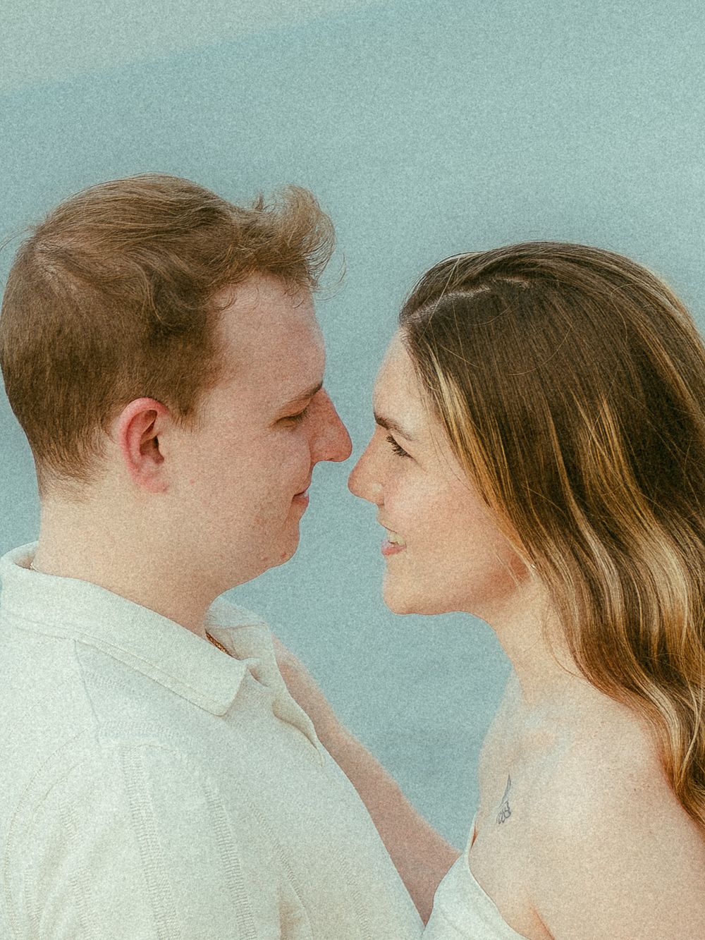 A romantic couple gazes into each other's eyes outdoors, with a soft blue sky background.