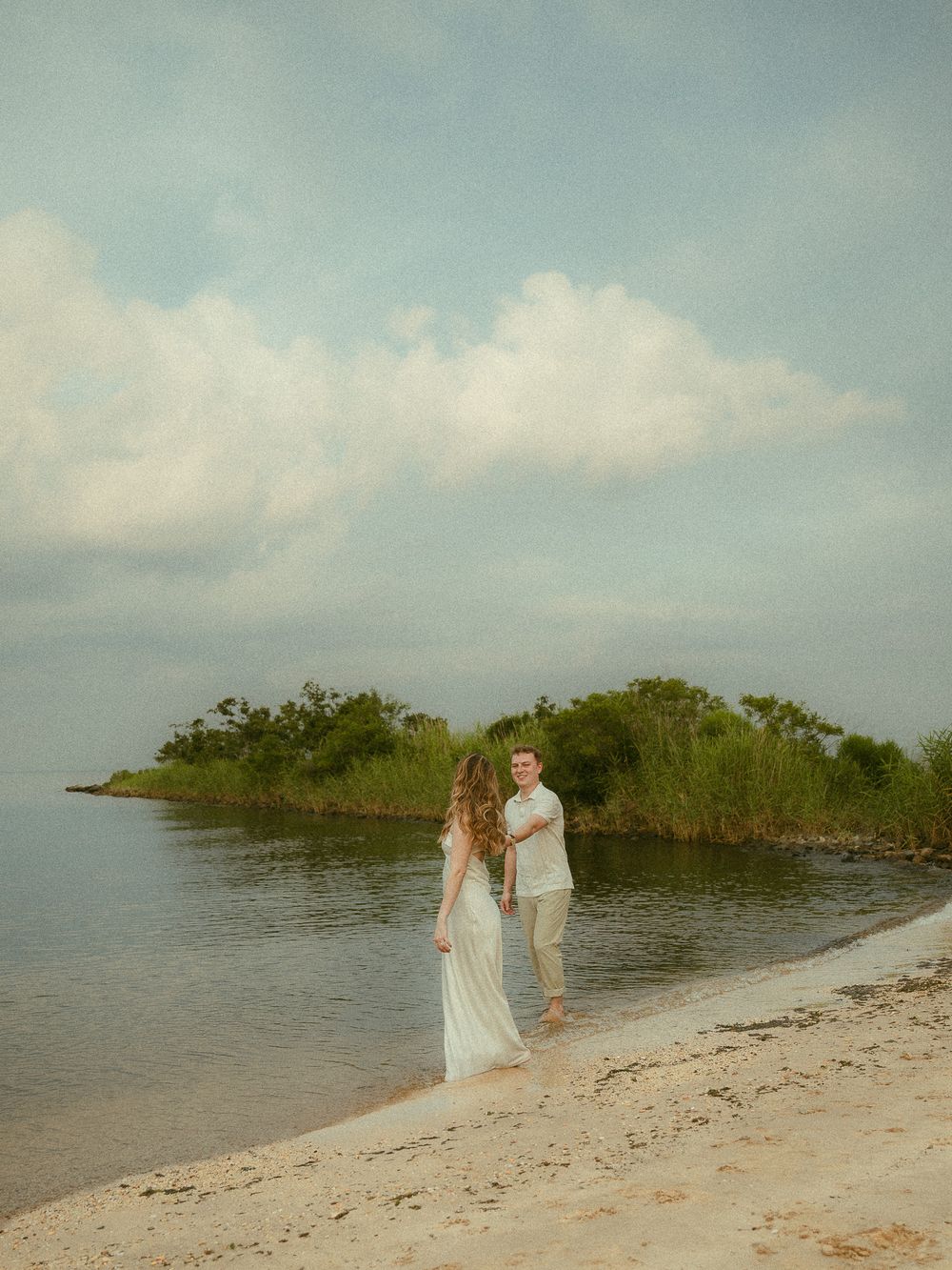 Couple holding hands on a sandy beach shore with lush green trees and cloudy sky backdrop.