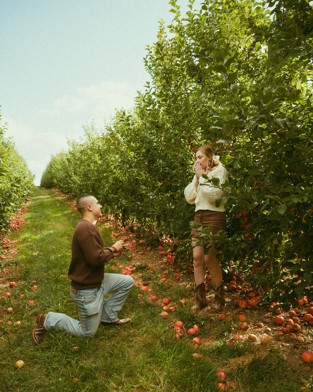 Person proposing on one knee in an apple orchard, partner covers mouth in surprise, red apples on ground.