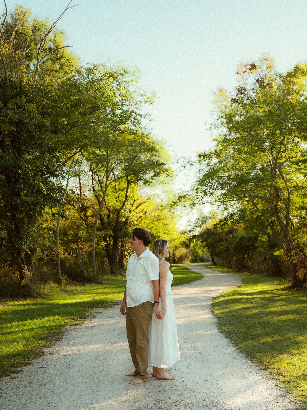 Couple standing face to face on a scenic gravel path surrounded by lush green trees in golden hour light.