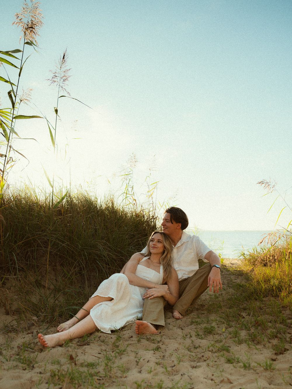 Couple sitting together on sandy beach dunes with sea oats and ocean in background at golden hour.
