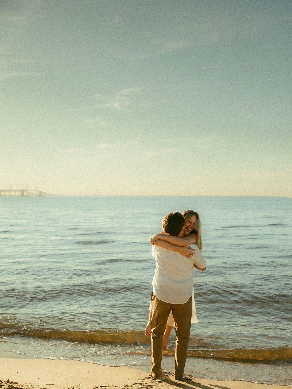 A couple embraces romantically on a sandy beach at sunset with calm ocean waters and a bridge in the background.