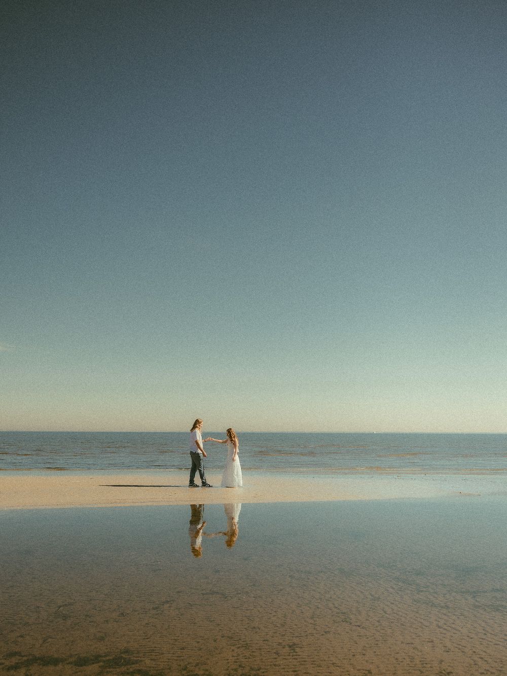 Couple holding hands on reflective wet sand beach with clear blue sky and calm ocean horizon.