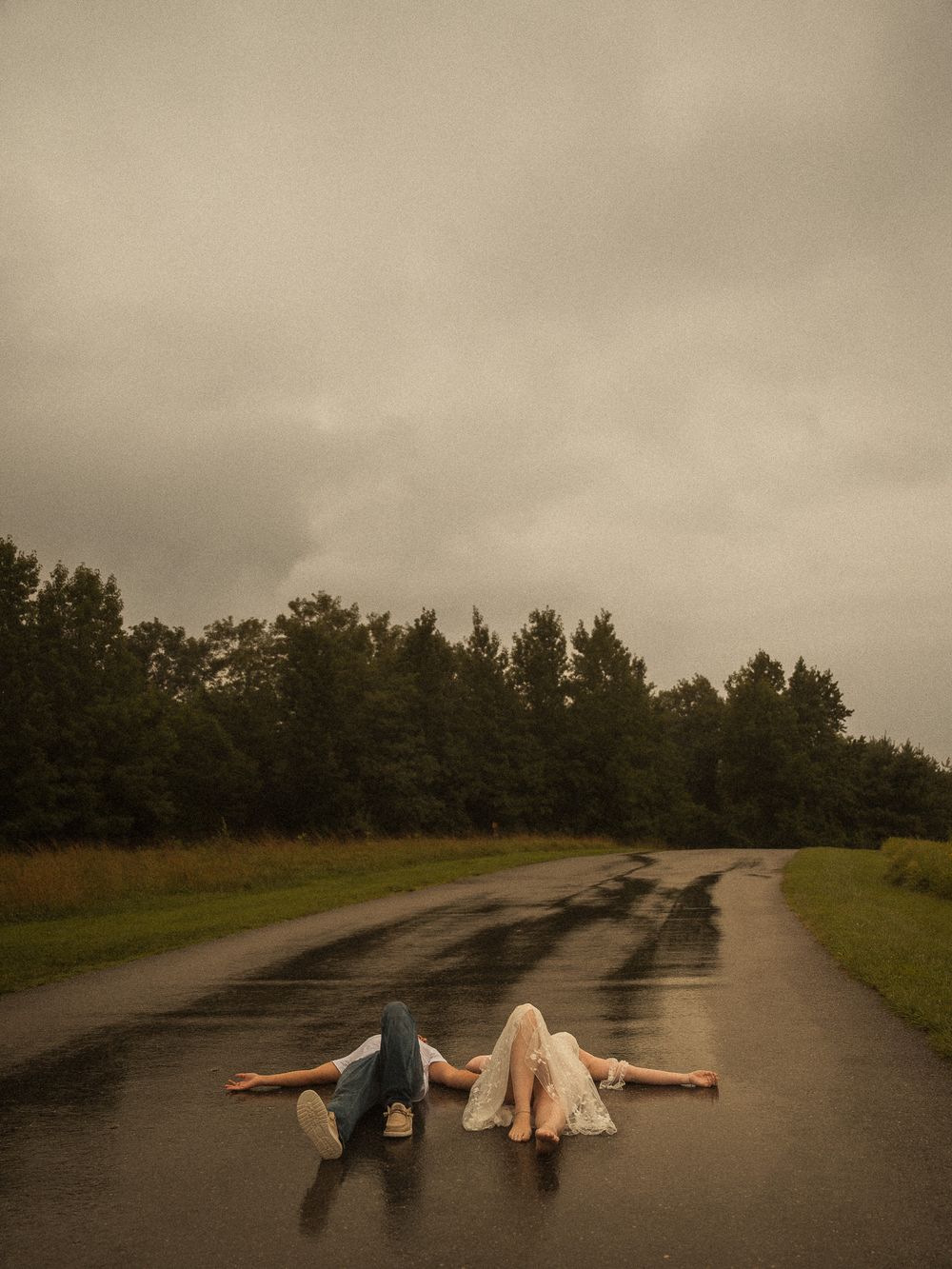 Two people lying on a wet road in the rain, surrounded by lush green trees under a moody overcast sky.
