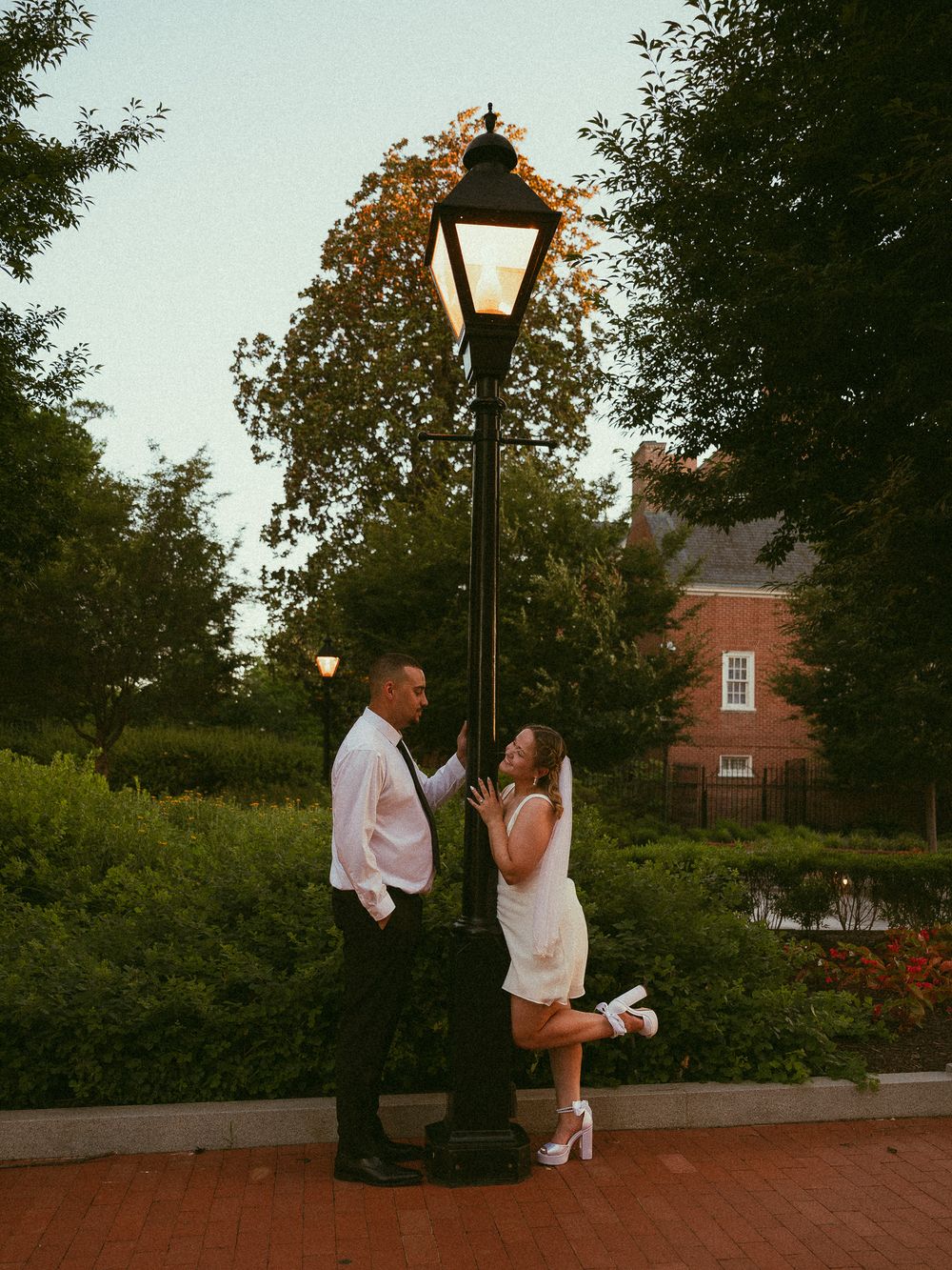 Couple posing by a glowing street lamp at dusk, woman in white dress and heels leaning playfully against the post.