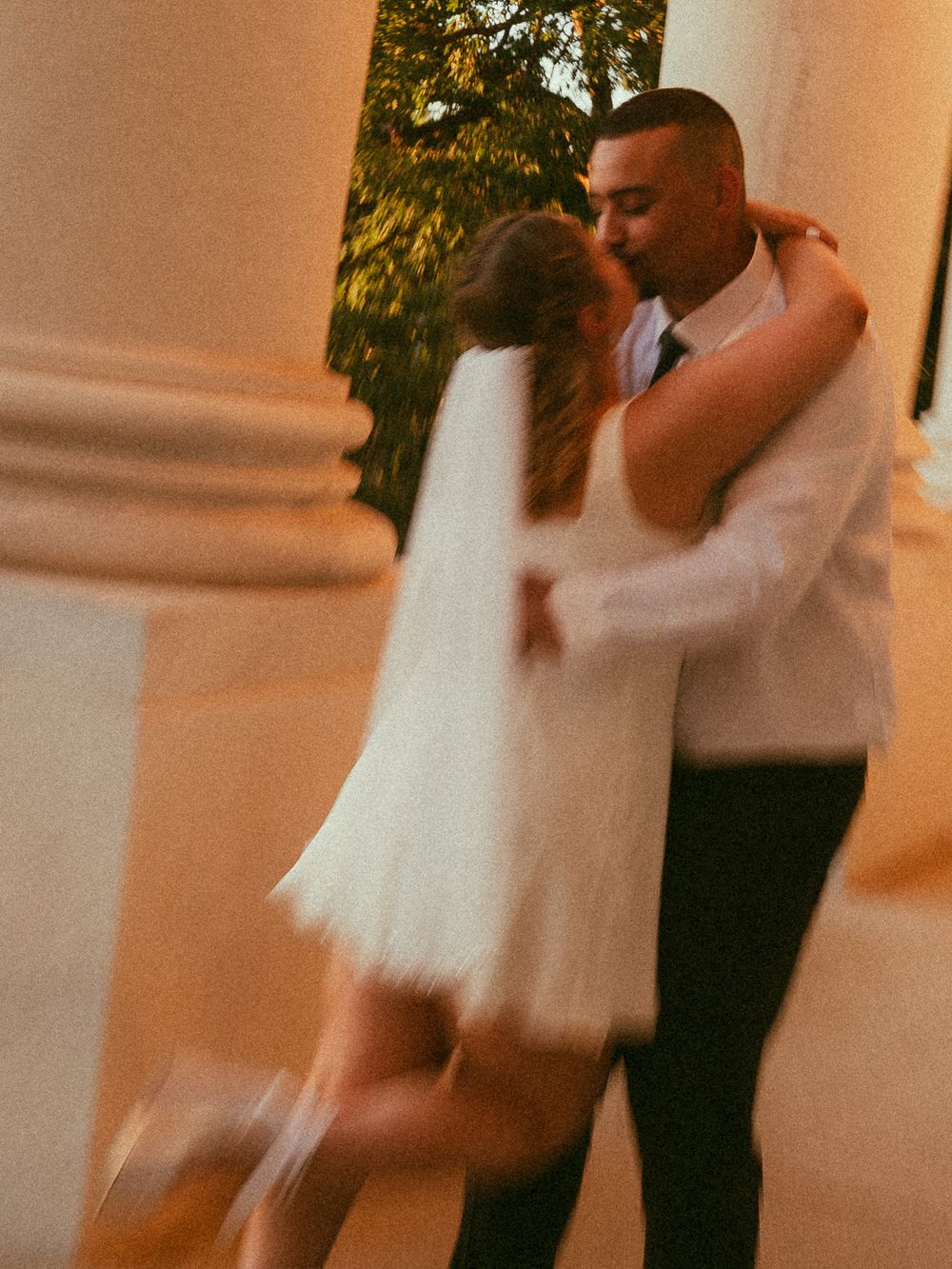 A couple shares a romantic first dance, bride in short white dress, groom in suit, at an elegant outdoor venue.