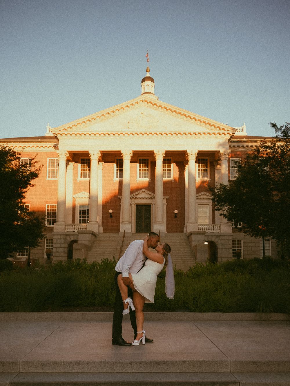 Couple sharing a romantic dip kiss in wedding attire in front of a grand columned building at golden hour.