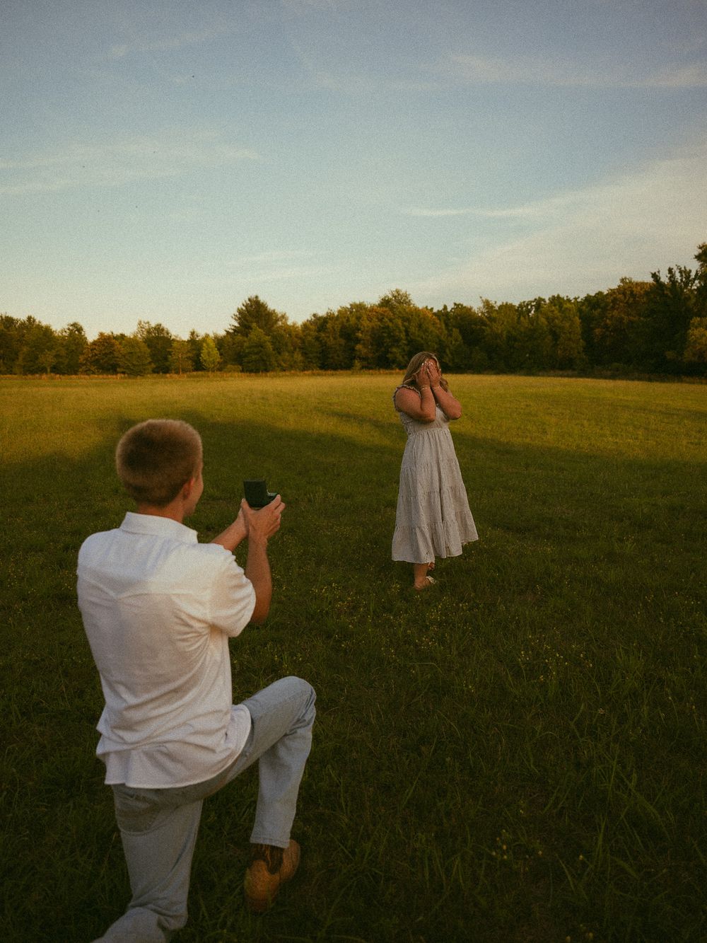 Man kneeling in green field proposing to woman at golden hour sunset outdoors.