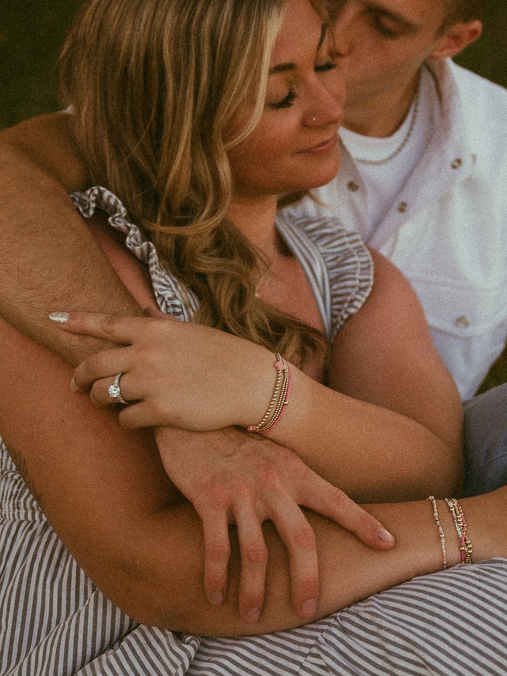 Couple embracing tenderly, woman wearing diamond engagement ring and gold bracelets, man in white shirt kissing her head.