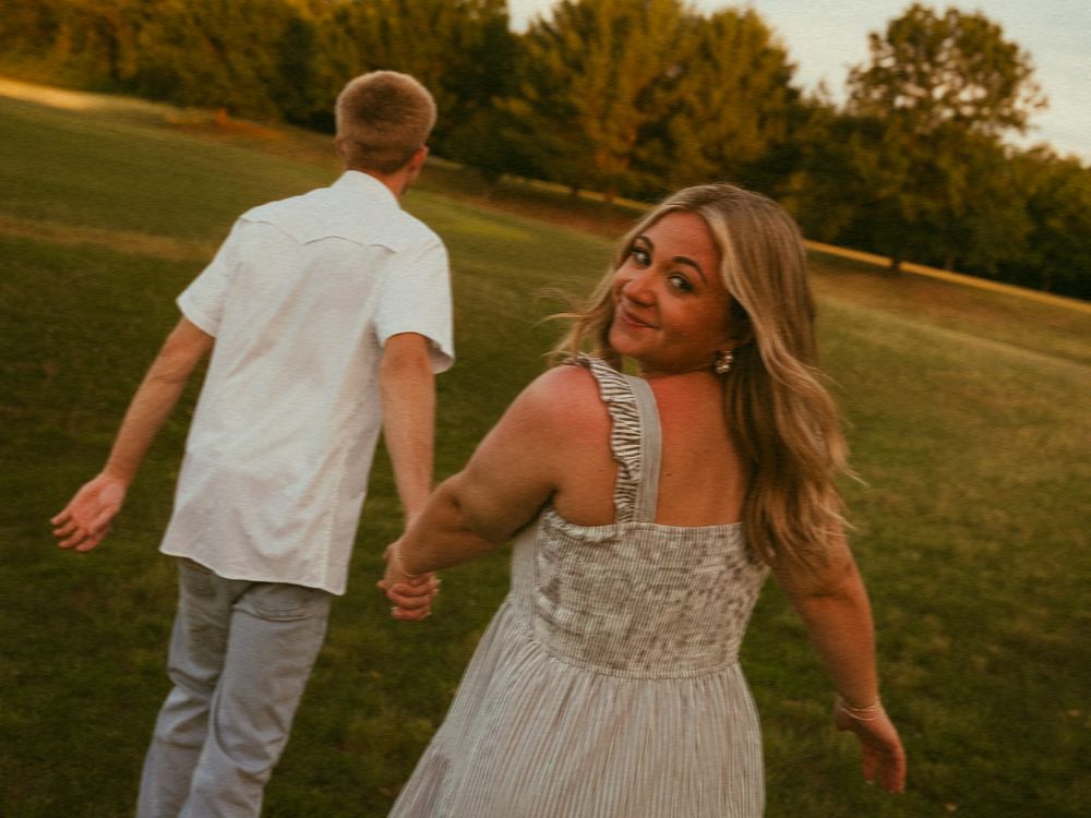 Couple holding hands walking through a sunlit park at golden hour, woman in floral dress smiling back.