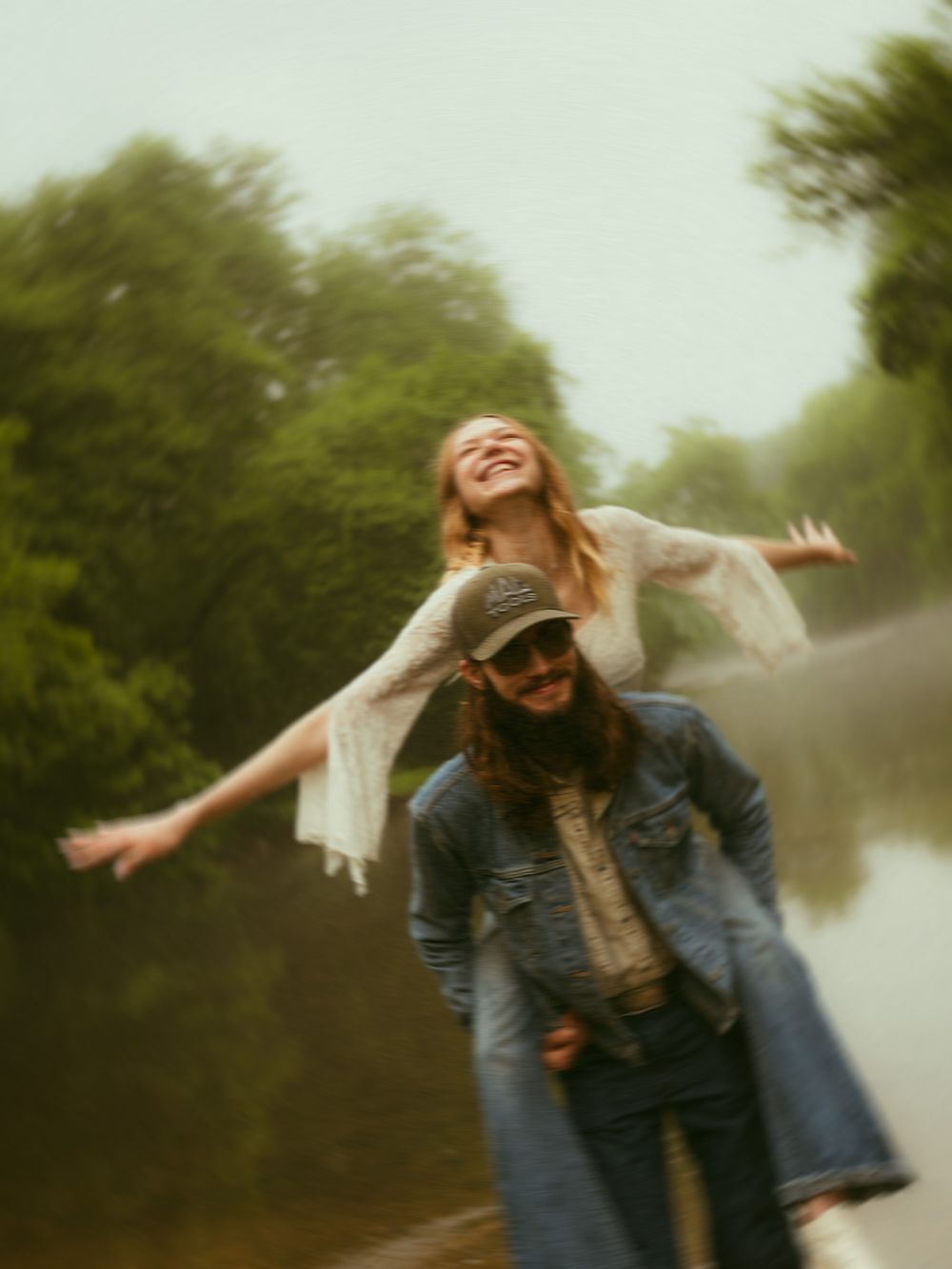 A joyful woman with arms outstretched rides on a man's shoulders in the rain near a lush green river.