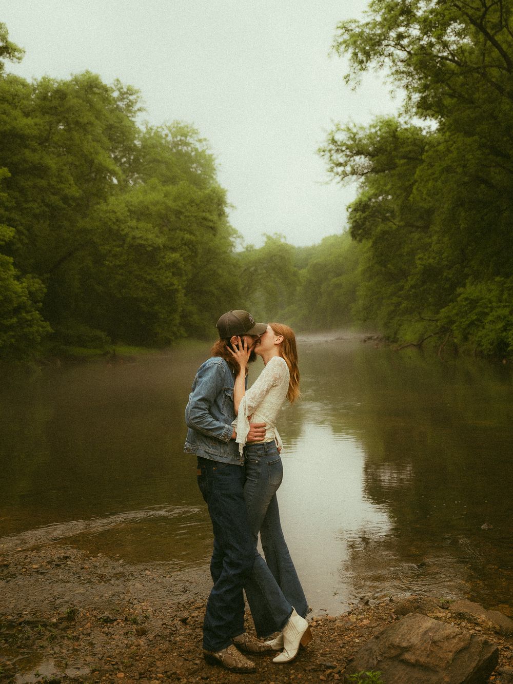 Couple sharing a romantic kiss by a misty river surrounded by lush green trees on a foggy day.