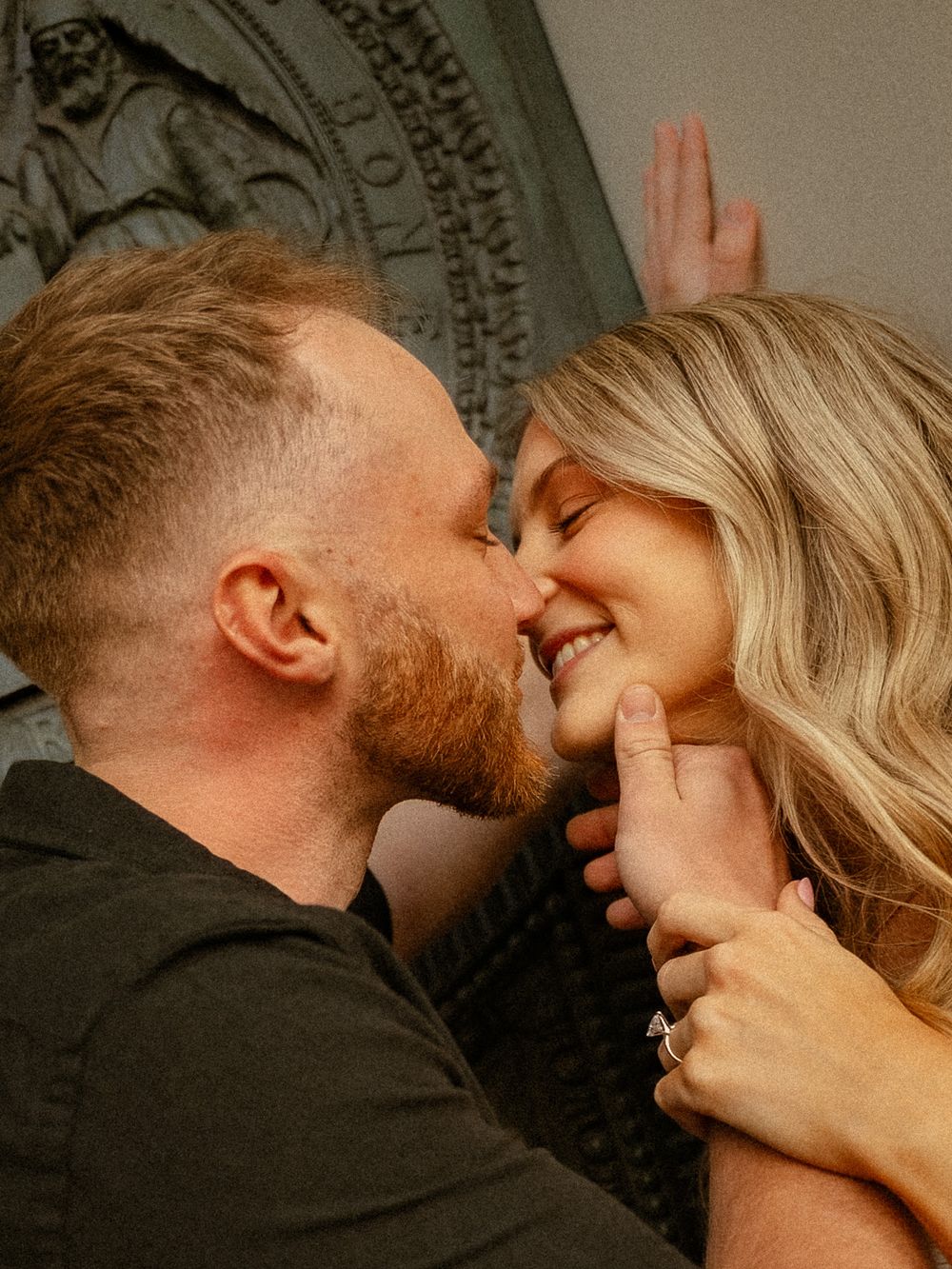A couple shares a romantic moment, nearly kissing, as the woman smiles wearing an engagement ring.
