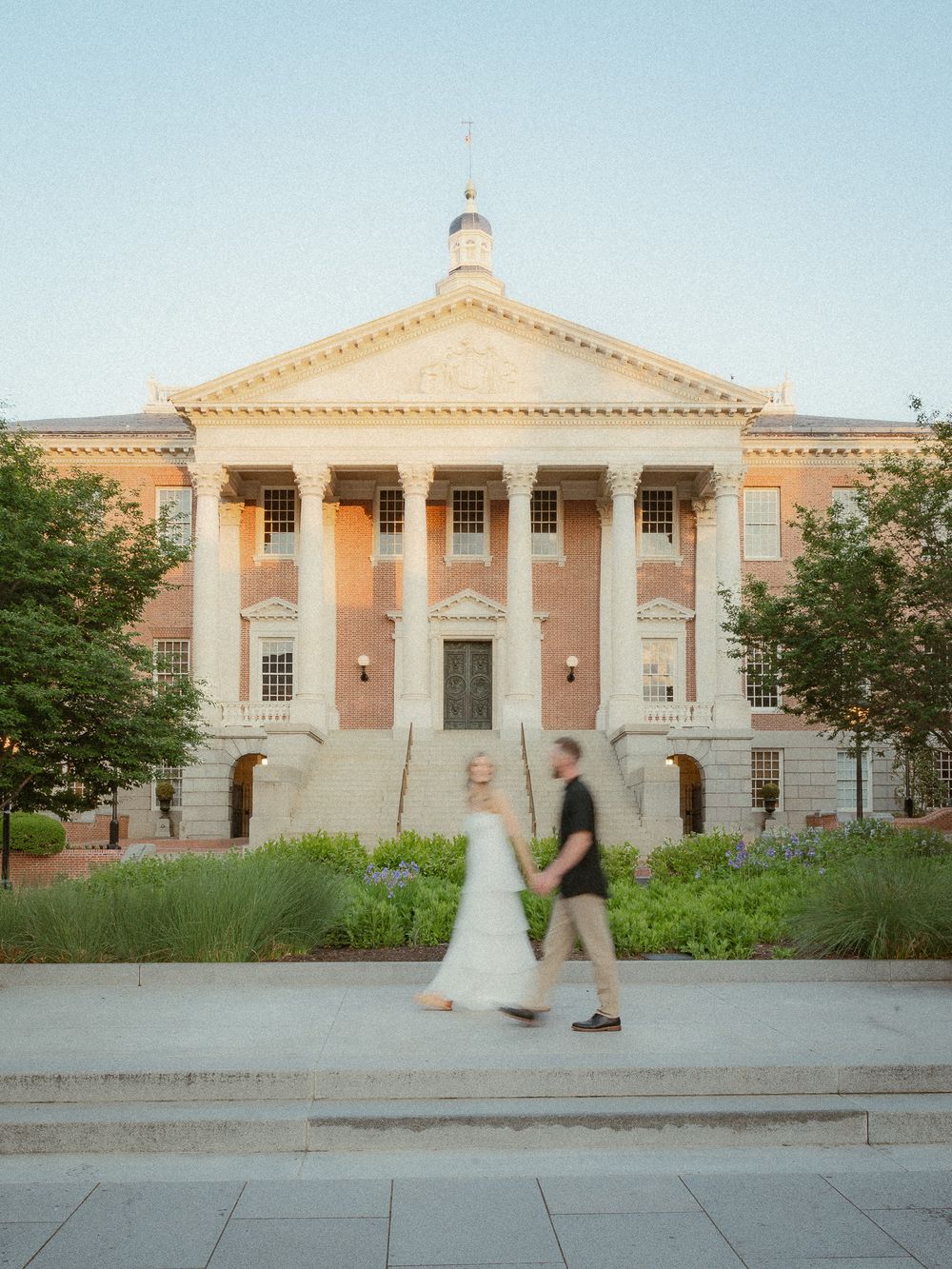 Couple walks past a grand neoclassical building with columns at golden hour, woman in white dress.