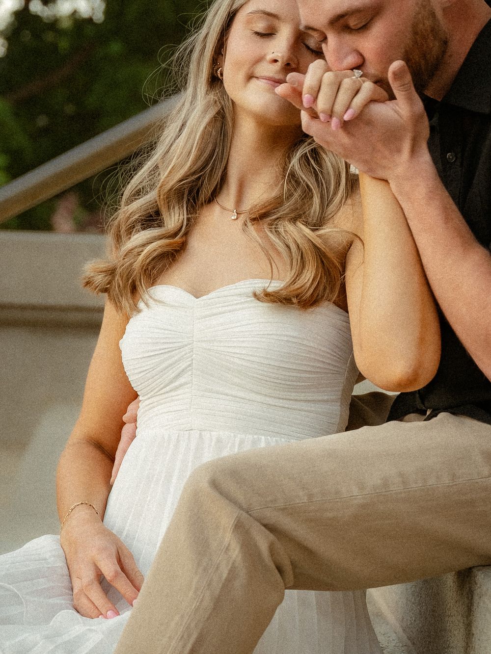 A bride in a white strapless gown smiles as her groom tenderly kisses her hand during an outdoor engagement session.