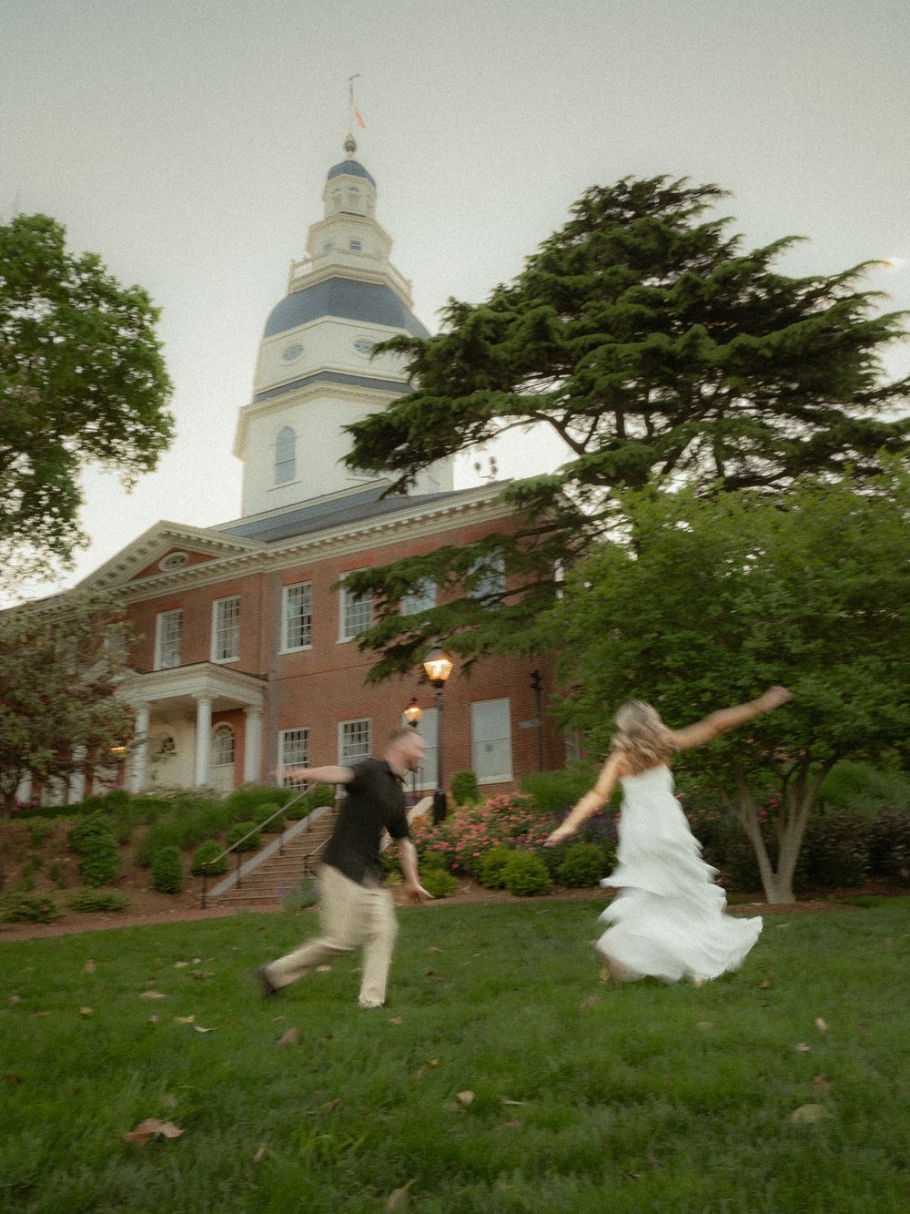 Couple in wedding attire running playfully on grass in front of a historic brick building with white dome at dusk.
