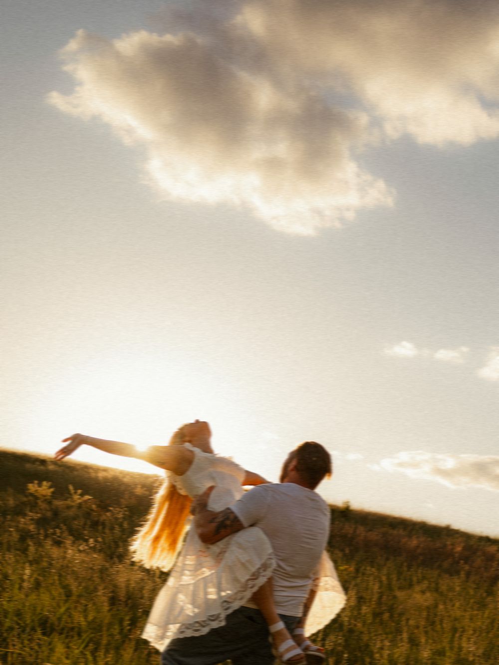 Couple dancing in golden sunset light in a green field, woman in white dress with long hair spinning joyfully.