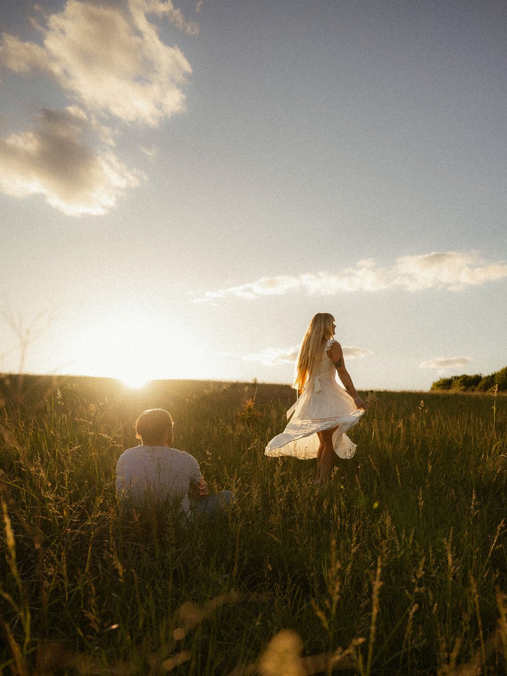 Couple in golden sunset field, woman in white dress standing while man sits in tall grass.