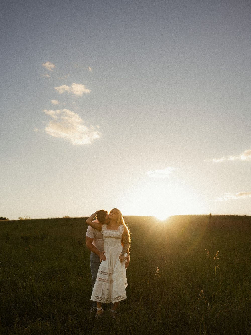 Couple sharing a romantic kiss at golden hour sunset in a lush green field, woman in white dress.