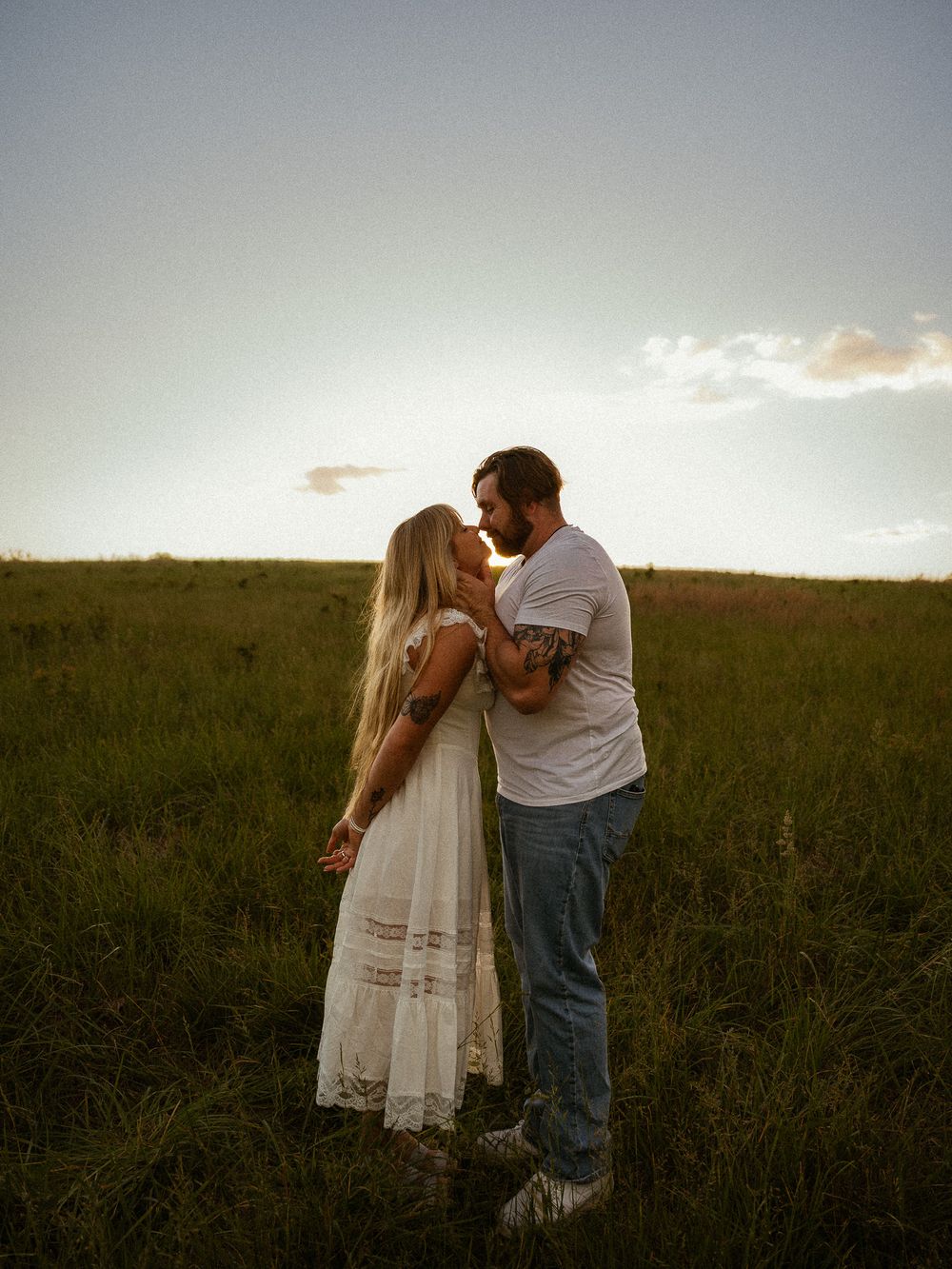 Couple sharing a romantic kiss at sunset in a lush green field, woman in white dress, man in jeans and white tee.