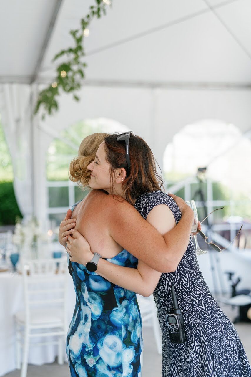Two women share a warm hug at an outdoor wedding reception under a white tent.
