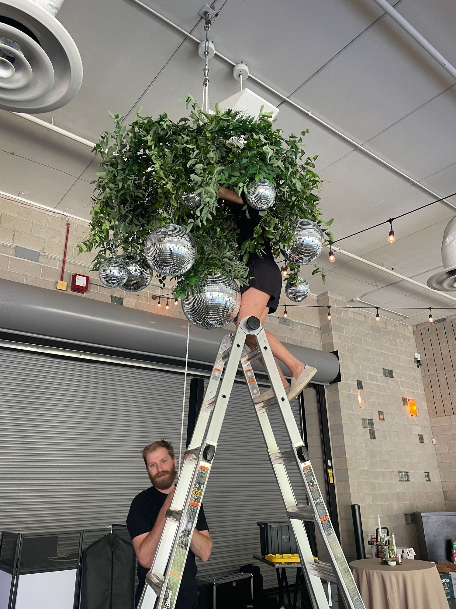 Two men on a ladder install a lush green plant chandelier with silver disco balls in an industrial venue.