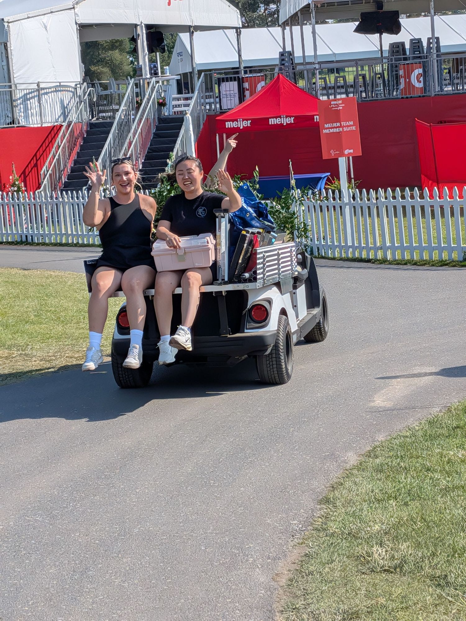 Two women riding on the back of a golf cart at a sunny outdoor event, smiling and waving near a Meijer tent.