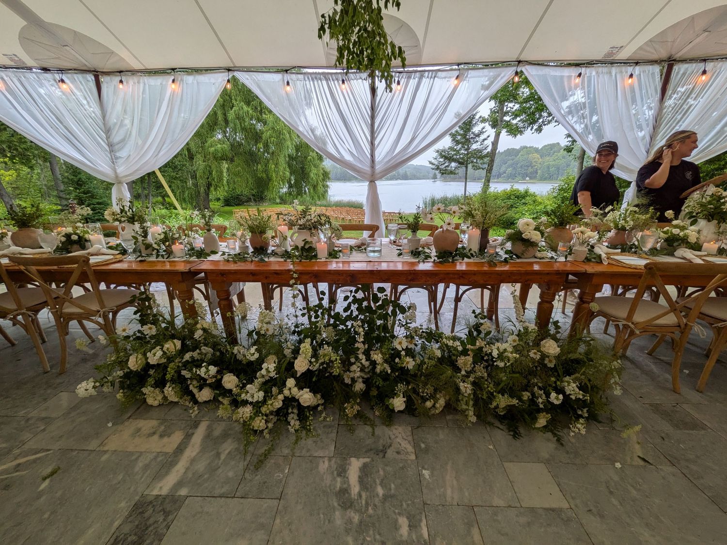 Elegant outdoor wedding reception table adorned with lush white floral arrangements under a draped tent overlooking a serene lake.