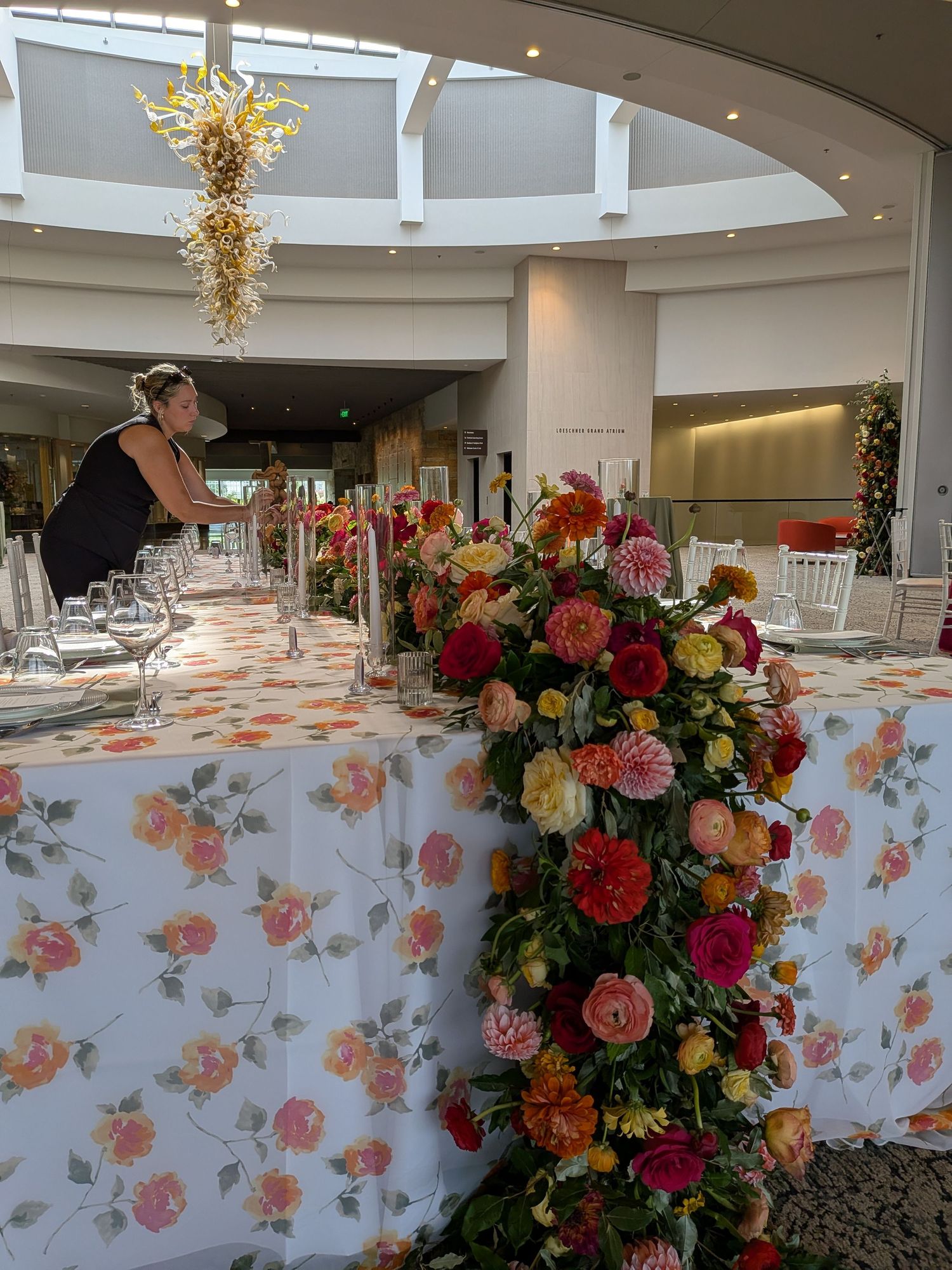 Elegant wedding reception table with floral tablecloth and cascading pink, orange, and yellow flower arrangement in a bright venue.