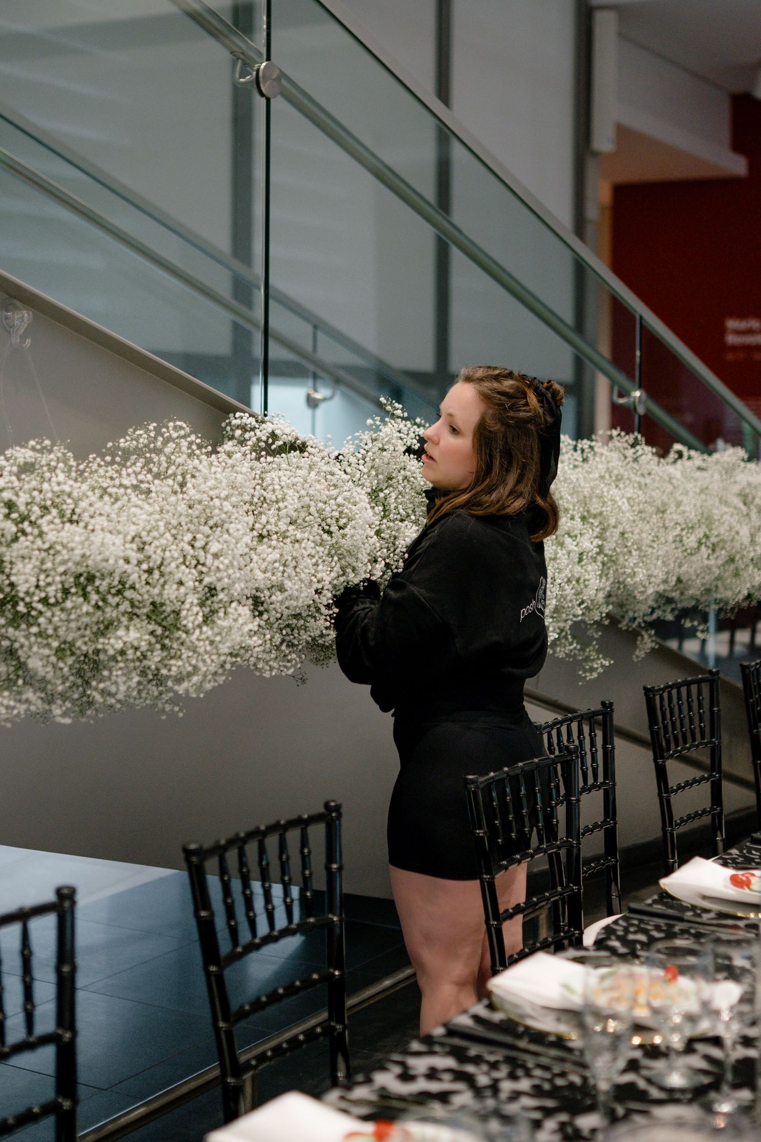 Woman in black holds white baby's breath flowers near black chairs at an elegant event setup.
