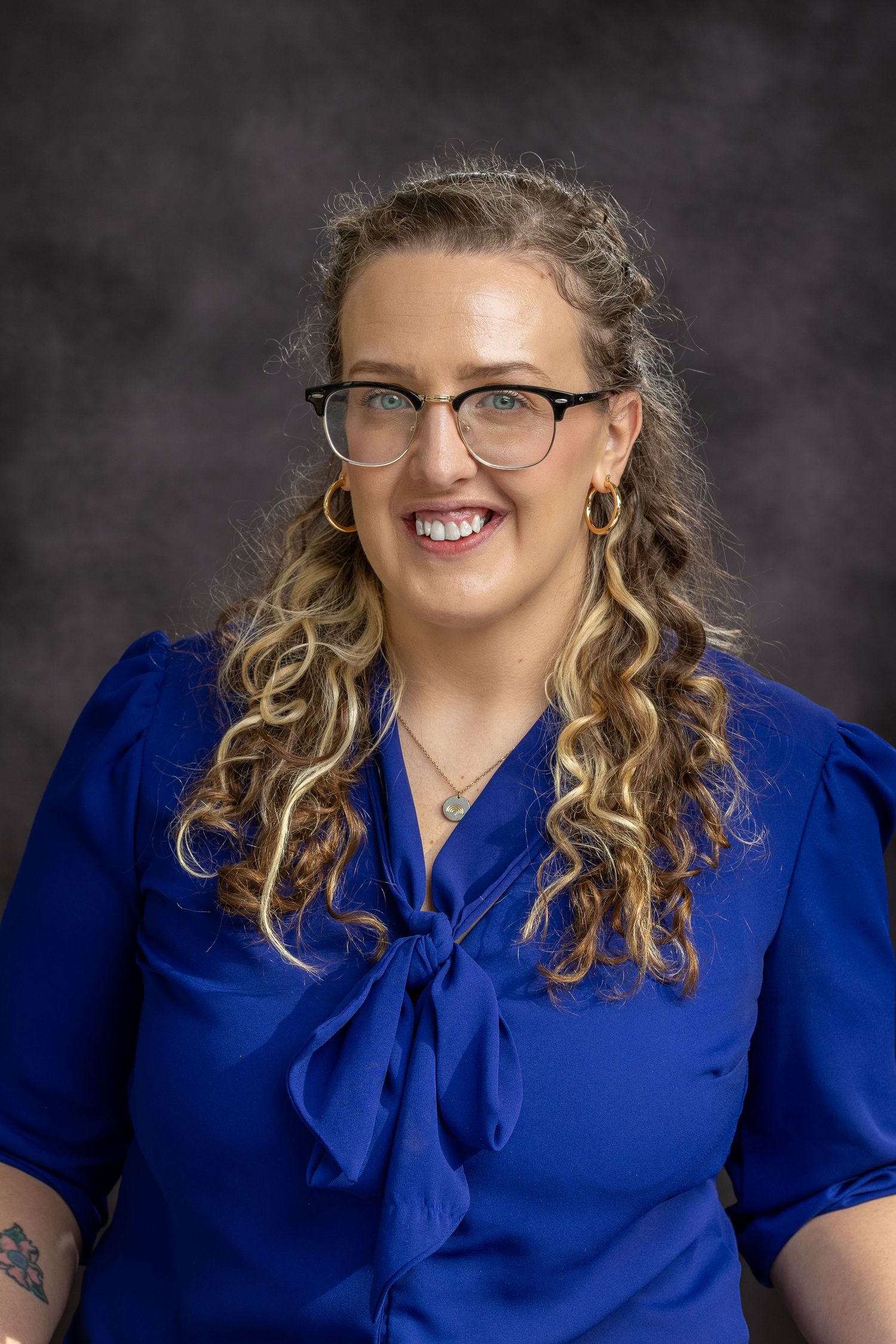 Professional headshot of someone wearing glasses and a royal blue blouse with a bow tie collar against a dark background.