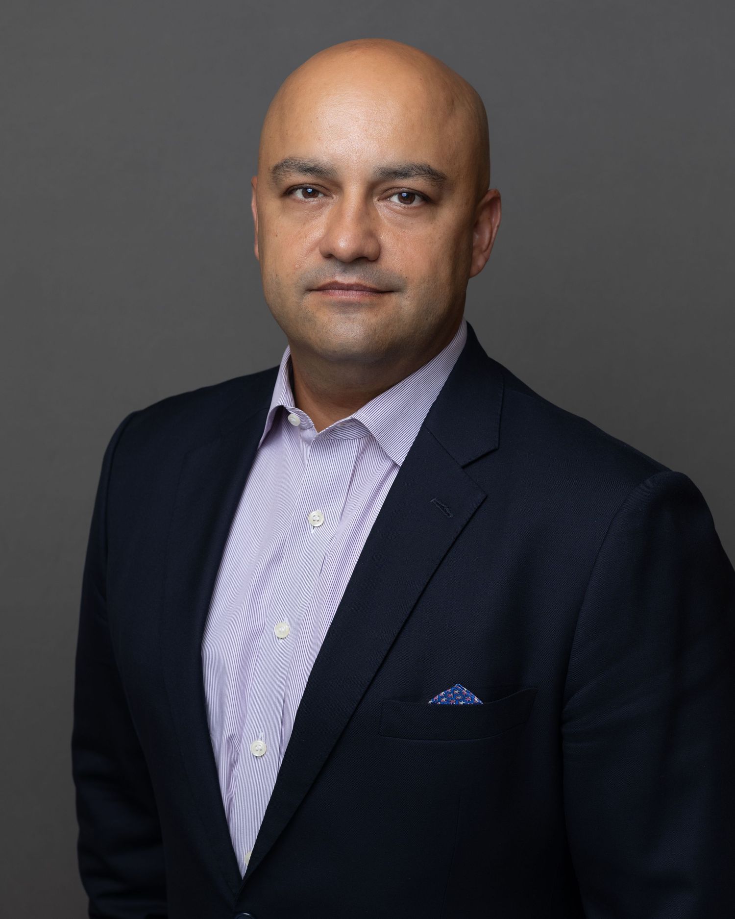 Professional headshot of person in dark suit and lavender shirt against gray background with confident expression.