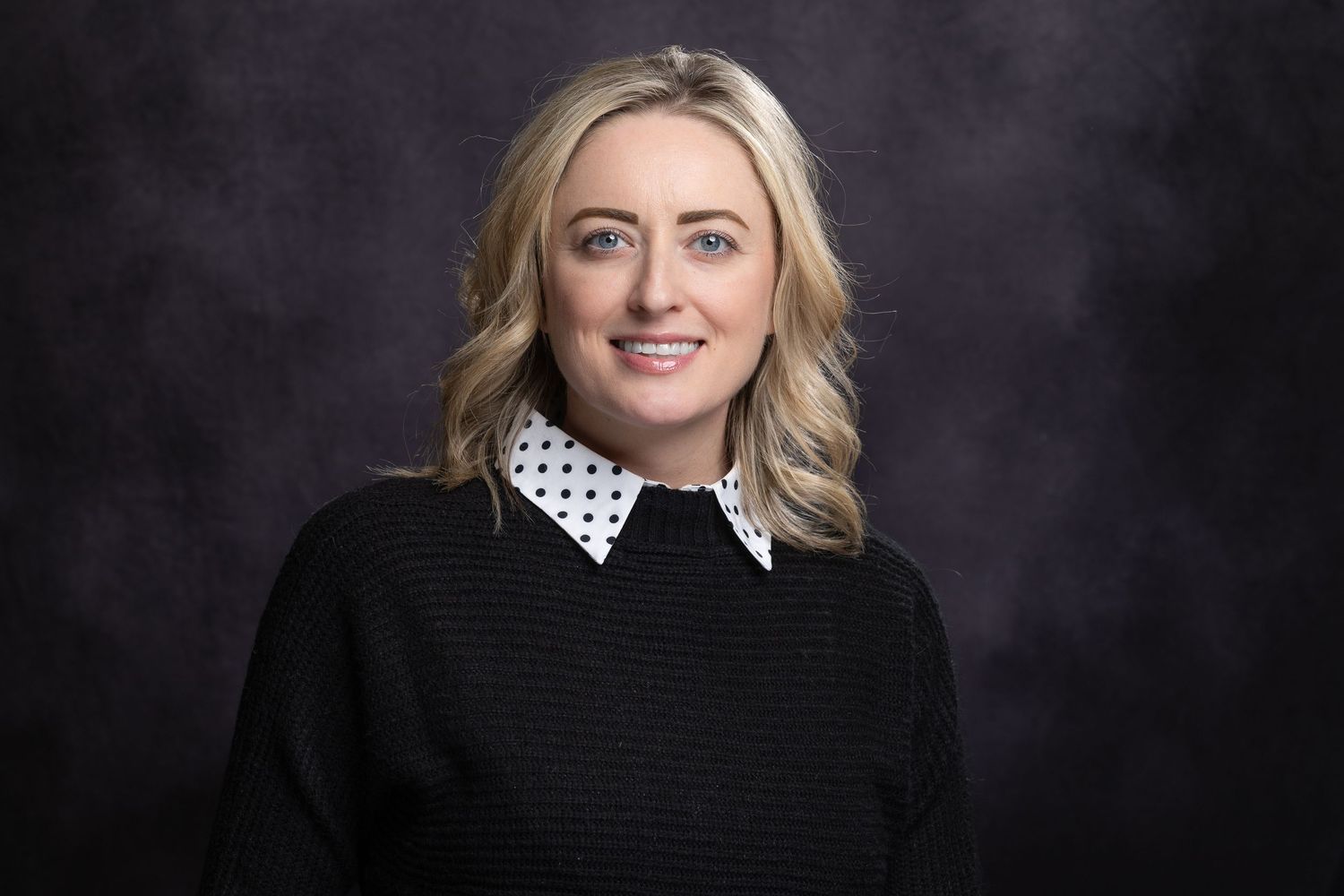 Professional headshot of a person wearing a black sweater with white polka dot collar against dark background.