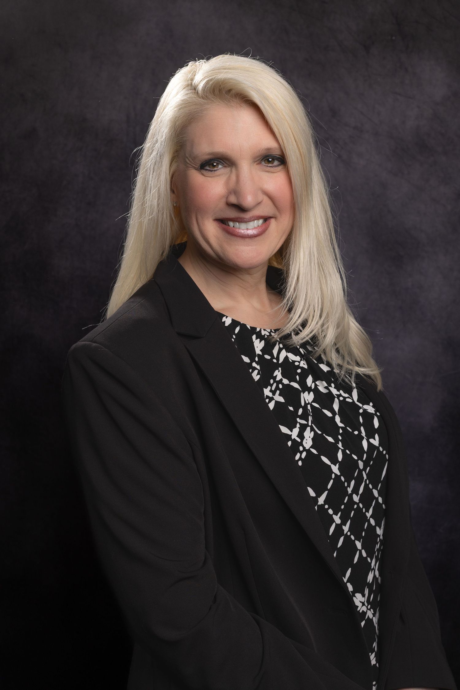 Professional headshot portrait with black blazer and patterned black and white top against dark background.