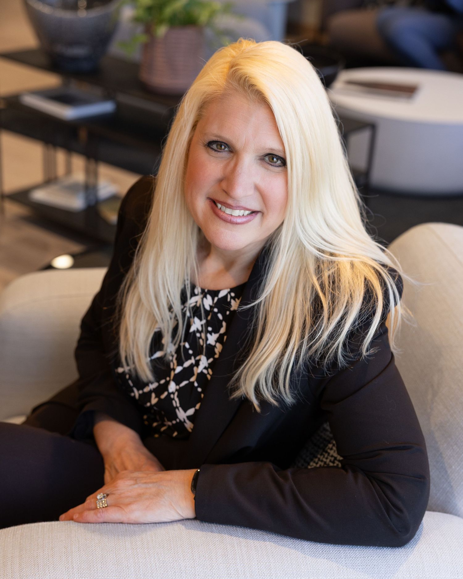 Professional headshot of someone with long blonde hair wearing a black blazer and patterned top seated on a couch in an office setting.
