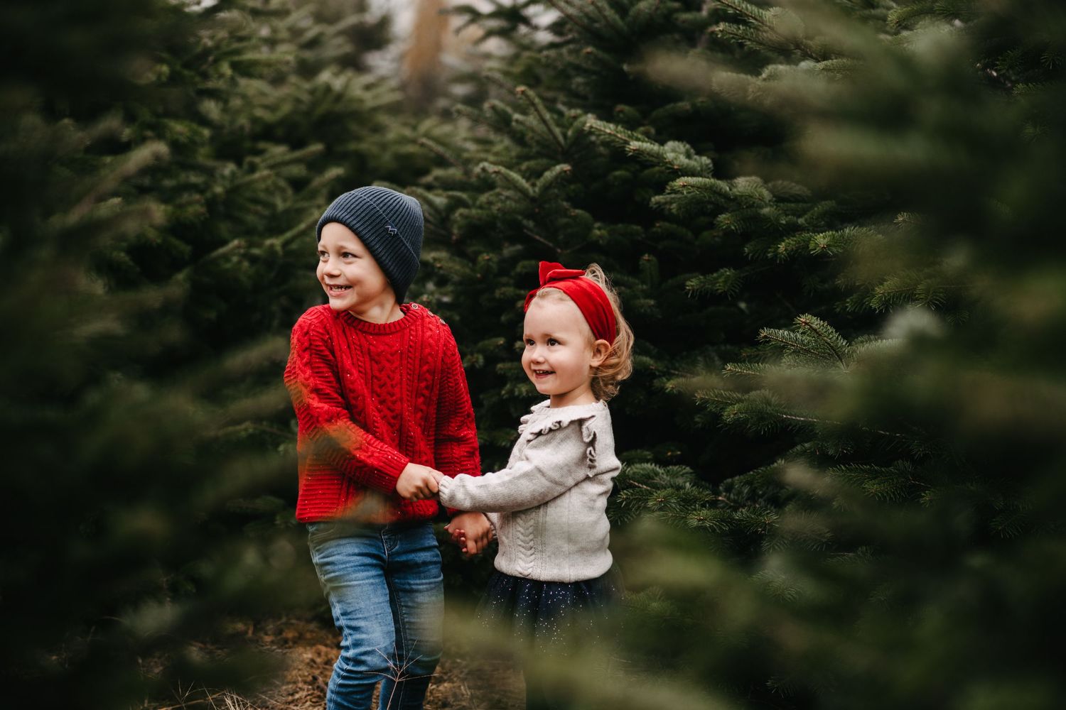 Two children in winter clothes holding hands and laughing together among evergreen trees on a misty day.