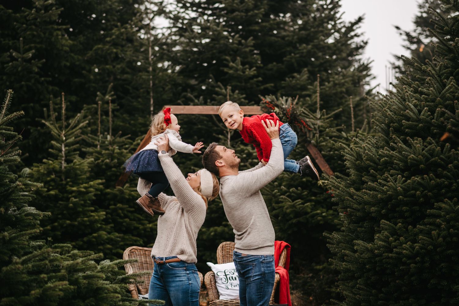 A couple lifts their children in front of evergreen Christmas trees during a family photo session.
