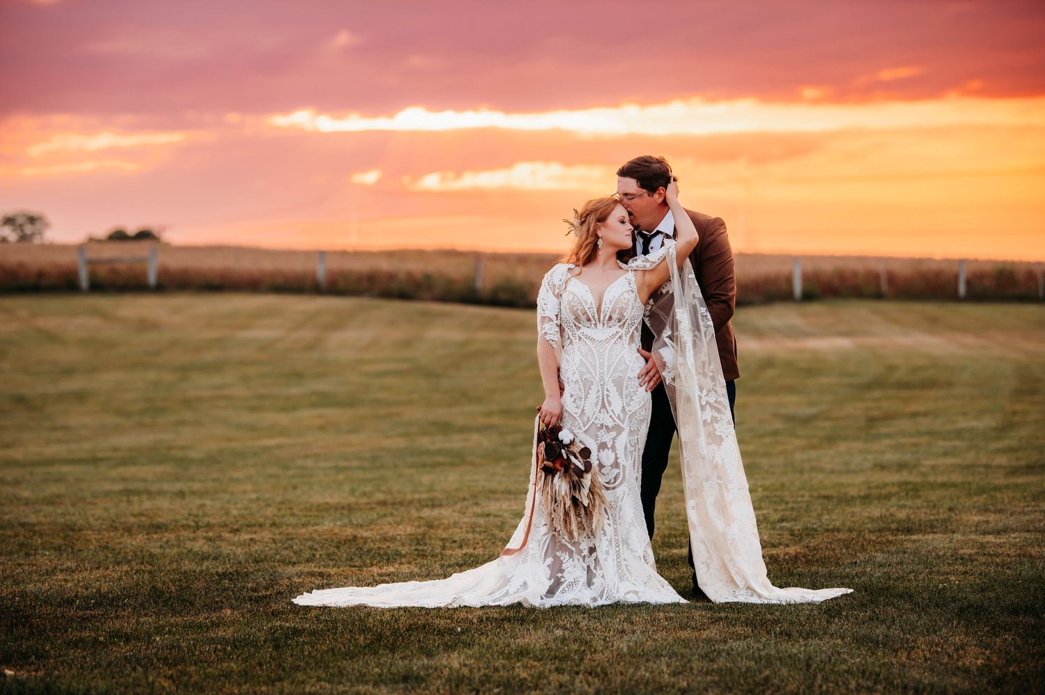A couple in lace wedding attire embraces in a field during a dramatic sunset.