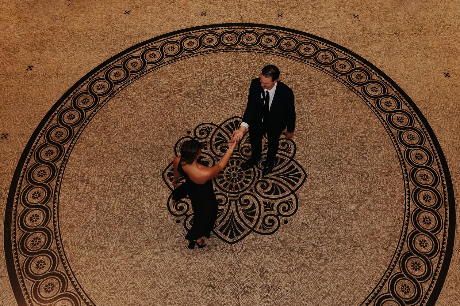 Two figures stand together on an ornate circular patterned floor from an aerial view.