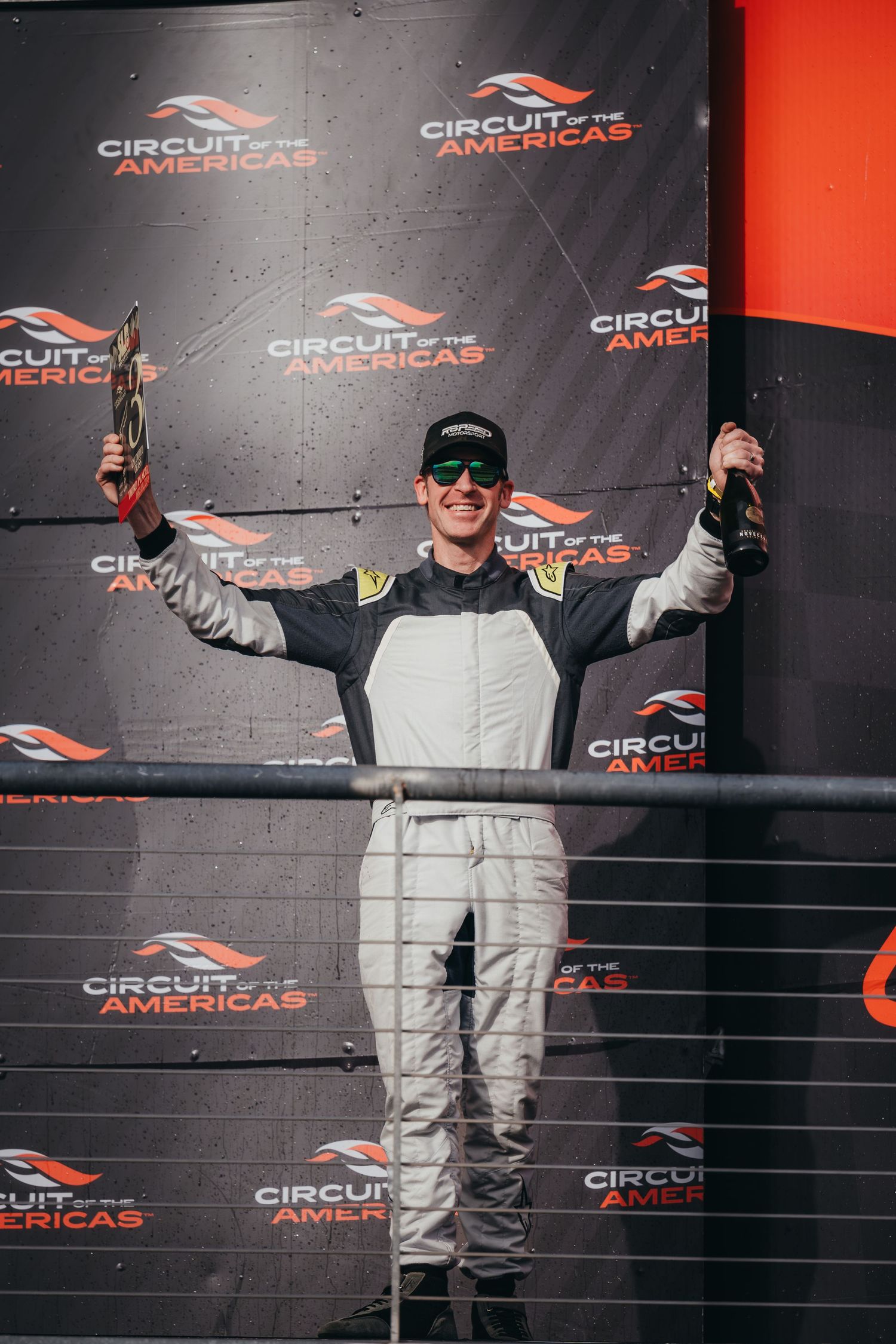 Racing podium celebration with drivers in white racing suits raising arms in victory.
