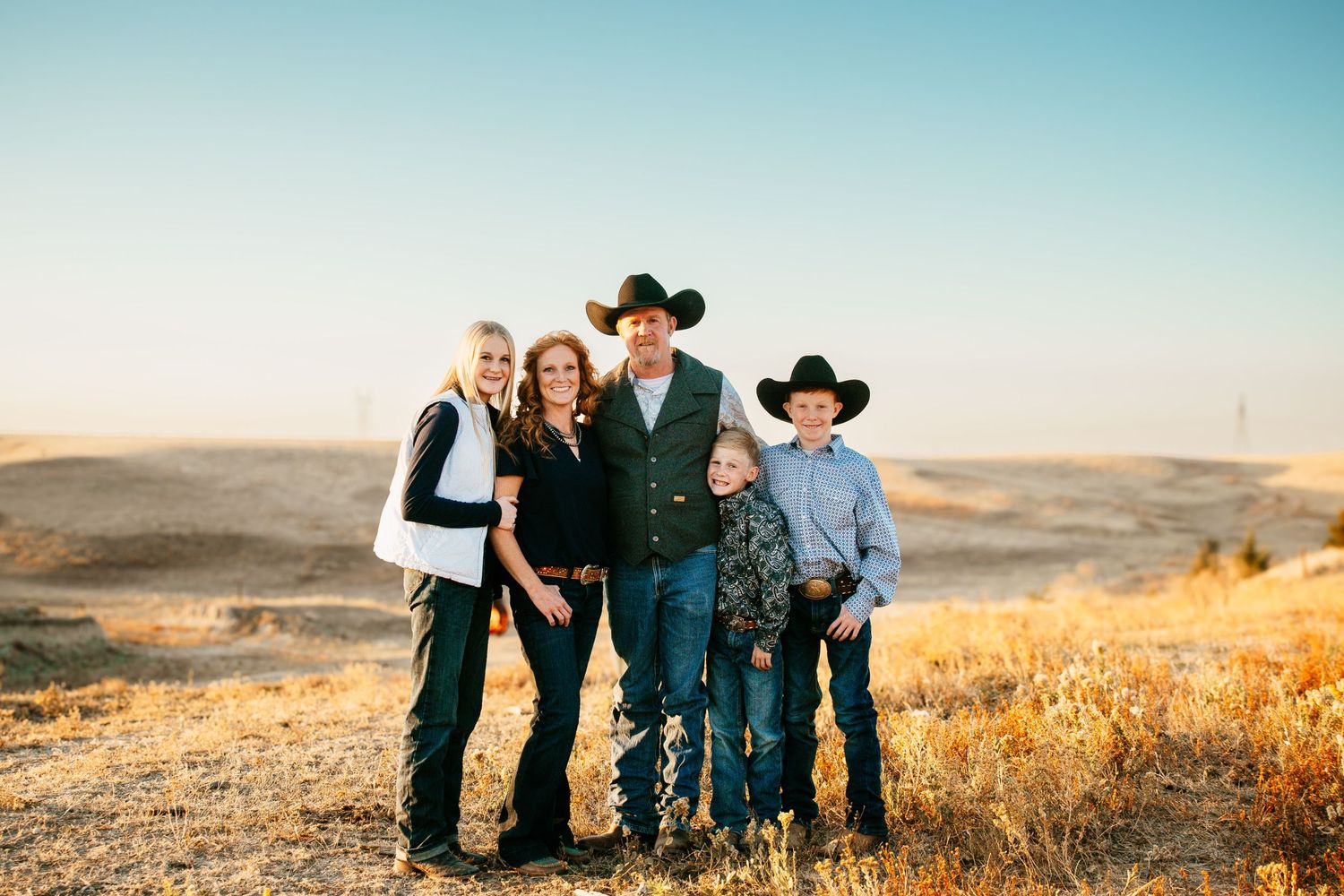 Western-styled group poses together against a scenic golden hillside backdrop at sunset.
