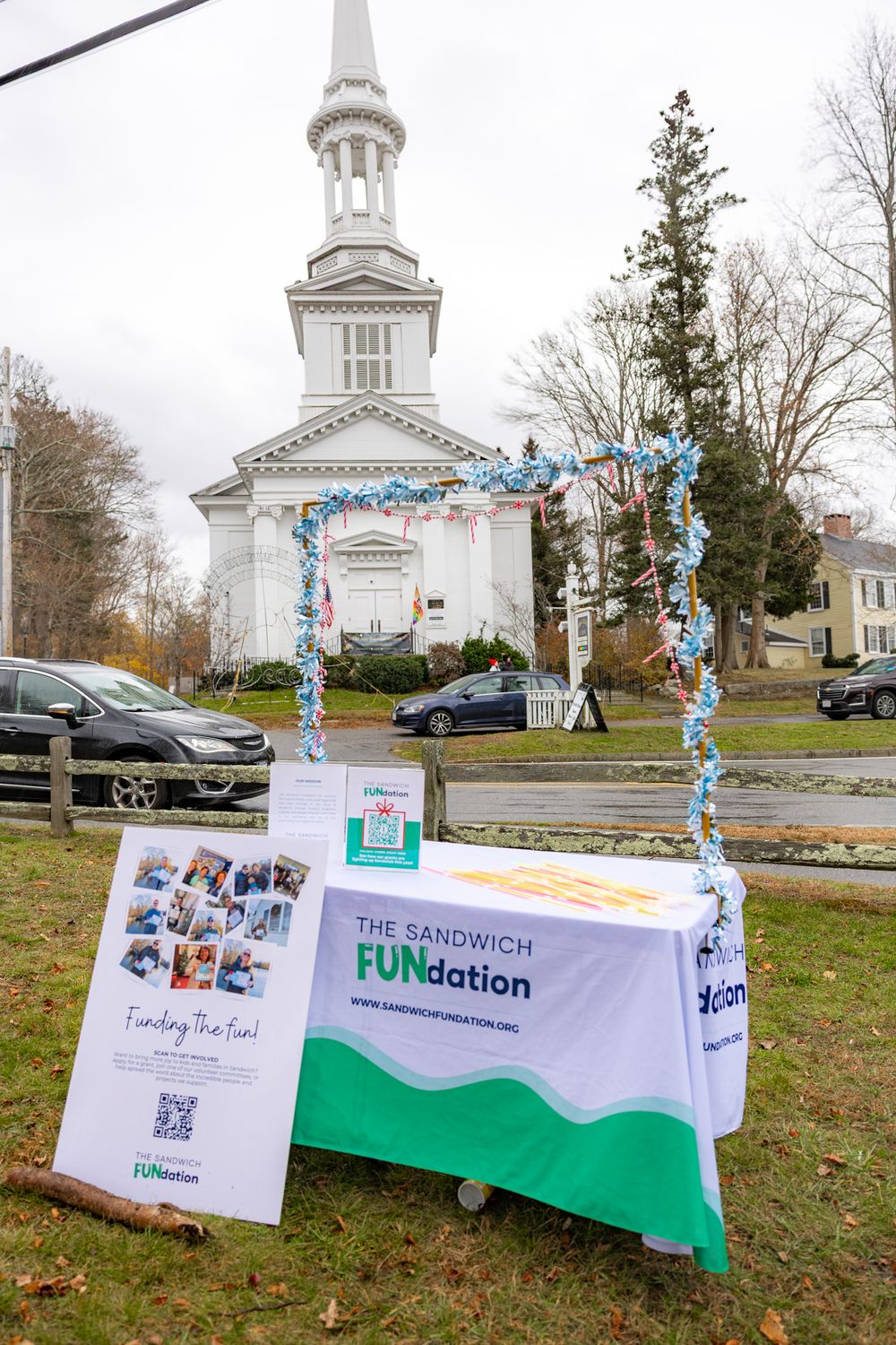 A white church building with a display booth featuring The Sandwich FUNdation banner and blue balloon arch decorations.
