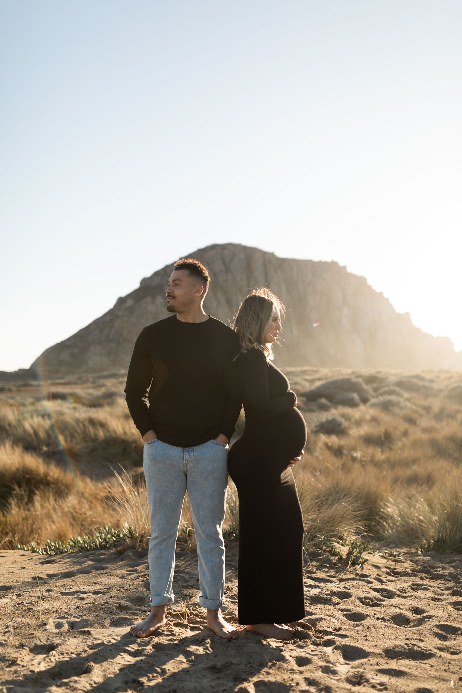 Couple standing on sandy beach with dramatic mountain silhouette at sunset.