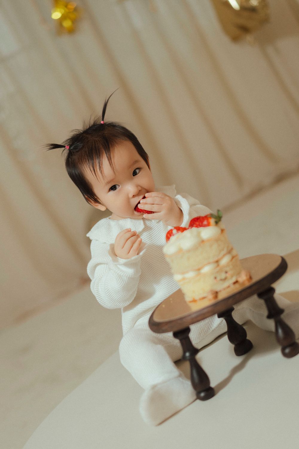 Baby in white outfit eagerly reaches for a tiered white cake with red decorations on a wooden cake stand.